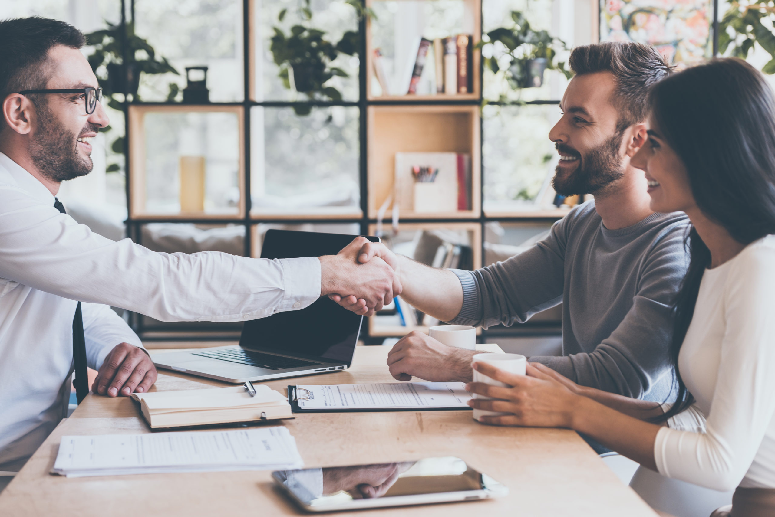 Deal! Side view of cheerful young man sitting close to his wife and shaking hand to man sitting in front of him at the desk