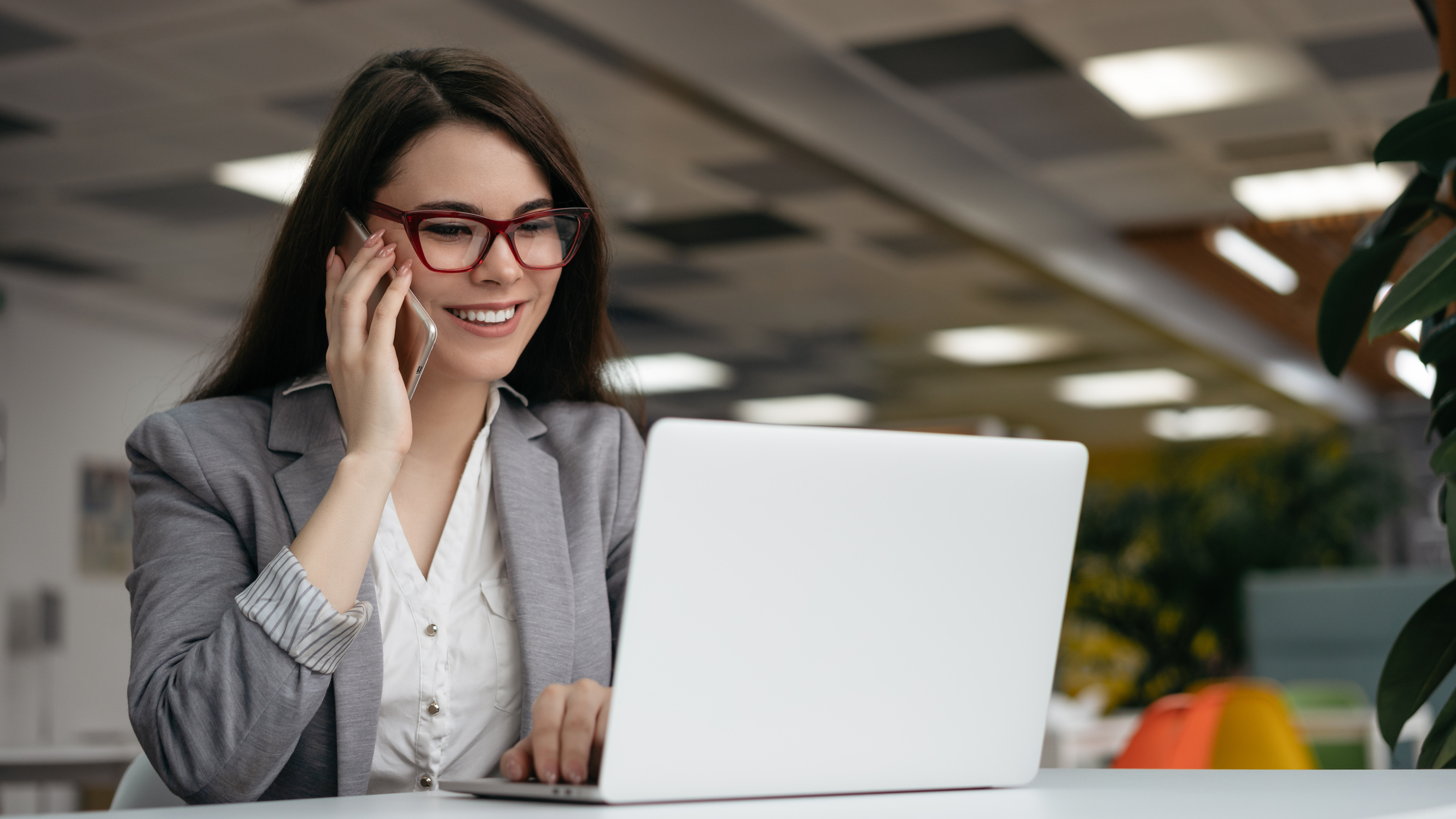 Businesswoman talking on mobile phone, working in office. Woman freelancer in stylish eyeglasses using laptop computer, looking at digital screen, communication at workplace