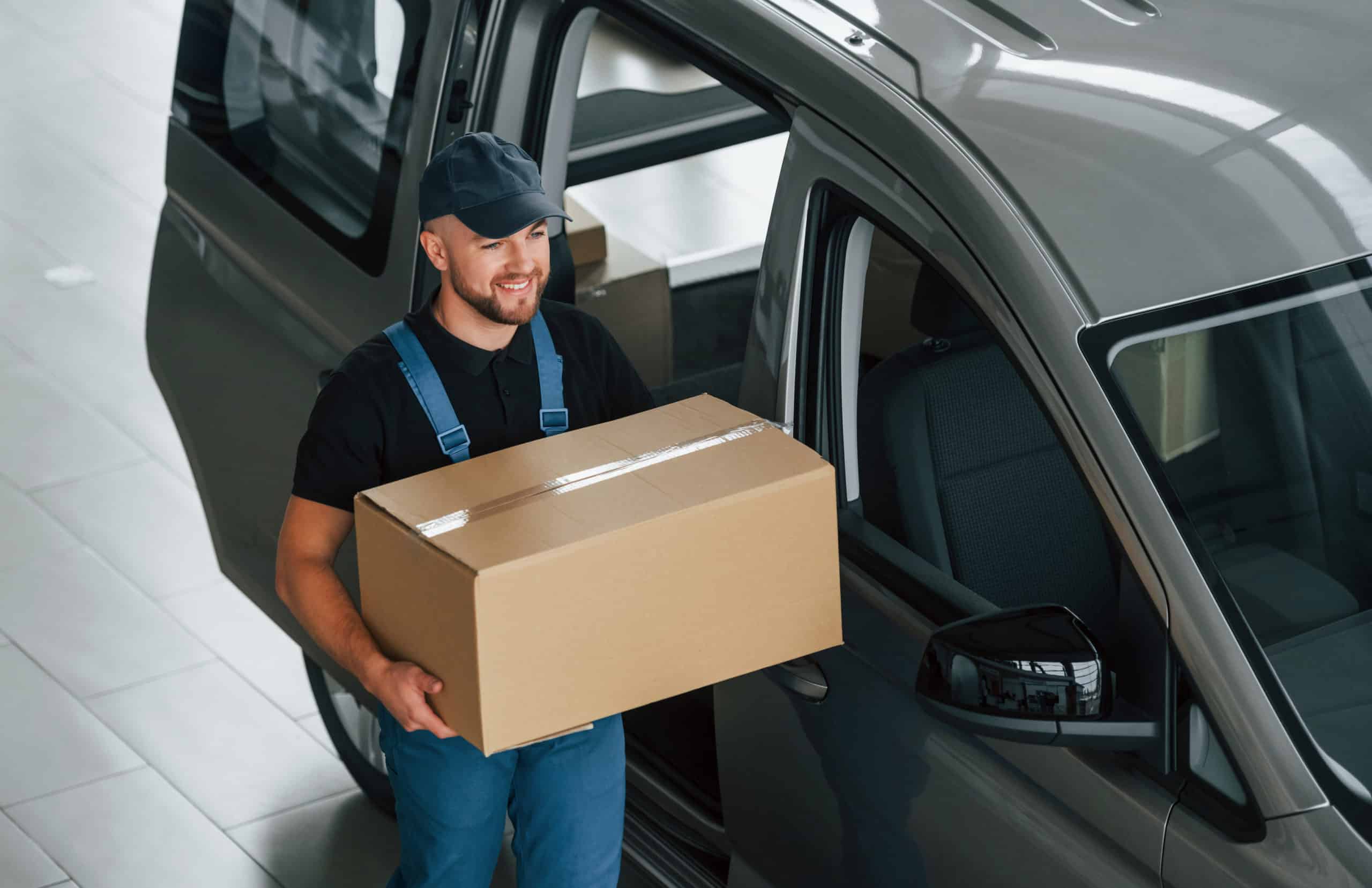 In blue colored clothes. Delivery man in uniform is indoors with car and with order