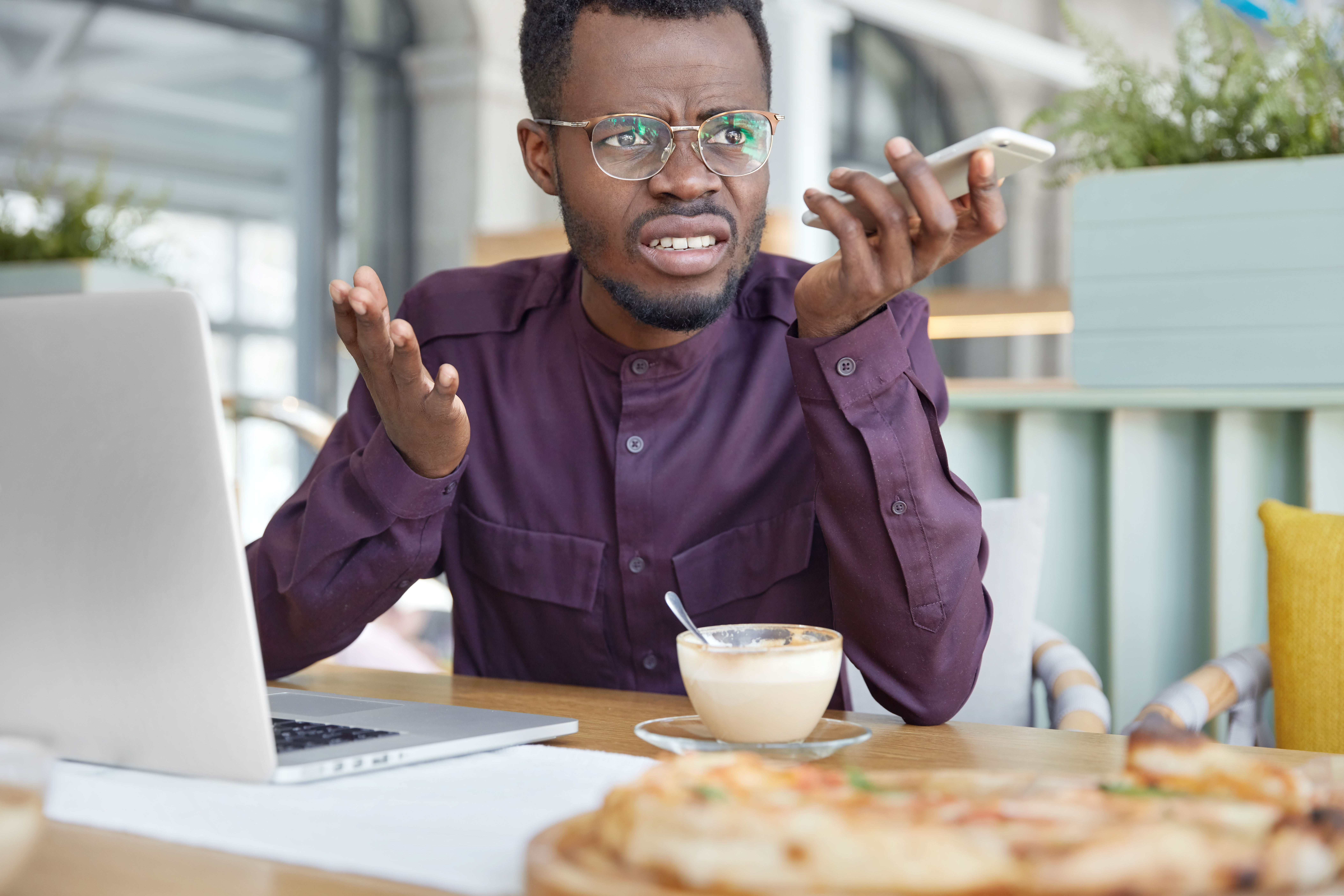 Irritated dark skinned young male enterpreneur being on working place feels very stressed and angry as can`t manage to do all work, surrounded with modern gadgets, looks in displeasure away.
